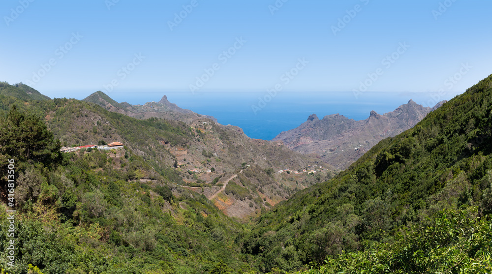 View from Tenerife's Anaga Mountains to the north over the Atlantic. Individual houses are built on the mountain slopes. There are some rocky peaks off the coast. It's a sunny and hazy autumn day.