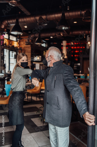 Senior businessman walks out of a restaurant. Young waitress kindly sees him off. They are wearing protective face mask as protection against virus pandemic.