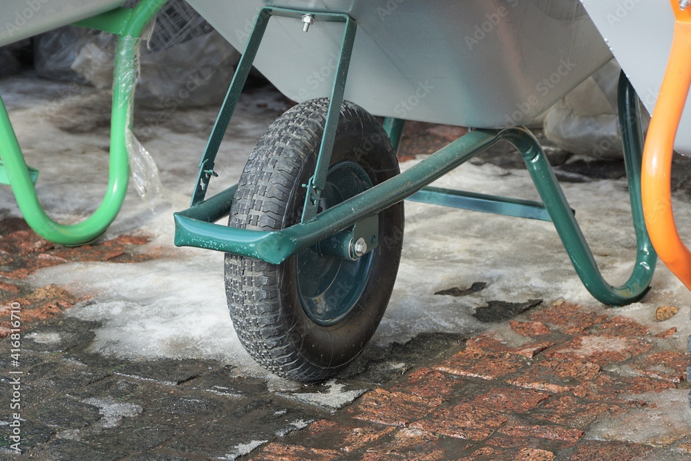 Fototapeta premium one black wheel on a green metal garden wheelbarrow stands on a brown sidewalk and white snow on a winter street