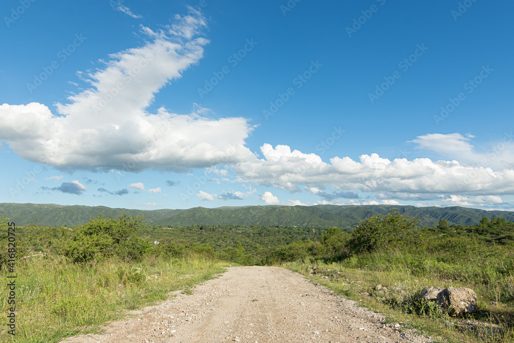 Horizontal landscape of a gravel road with mountains in the background, blue sky with major clouds