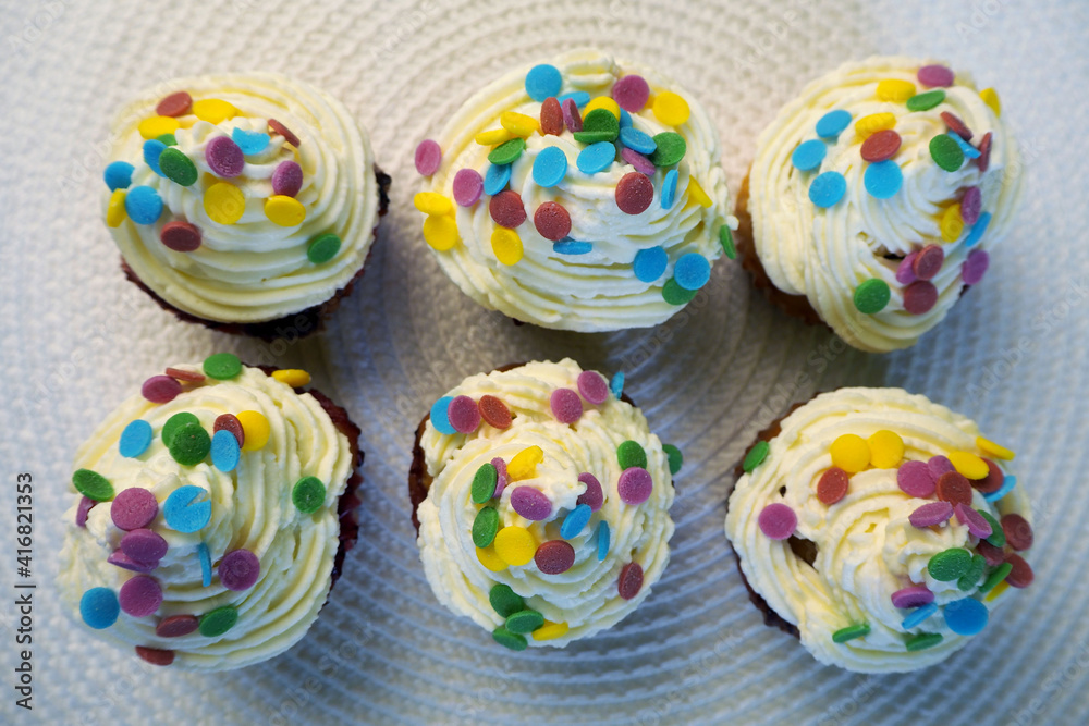 six yogurt muffins with red currants, spread with white cream of cream and mascarpone and decorated with colorful round splashes, lie on a white table stand with a view from above . homemade cakes for