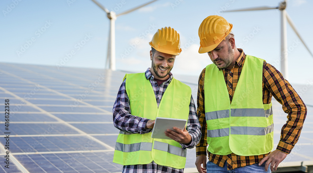 Men working at solar power station with digital tablet - Solar panels with wind turbines in background - Green energy renewable concept Men working at solar power station with digital tablet - Solar panels with wind turbines in background - Green energy renewable concept