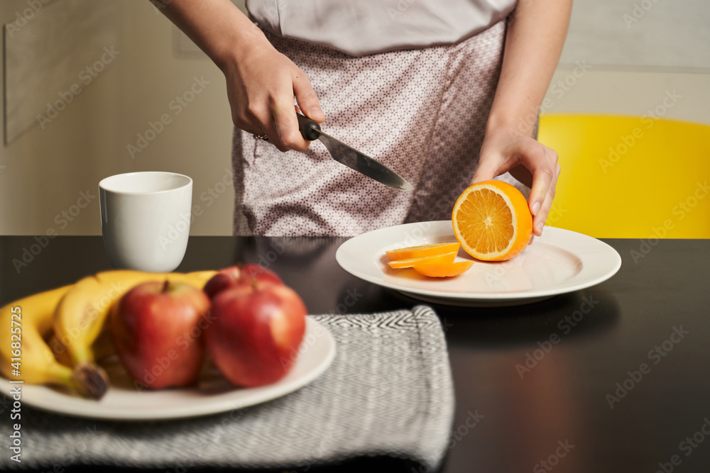 female hands cut an orange on the table. there are apples and bananas nearby and there is a cup. breakfast. healthy diet. selective focus