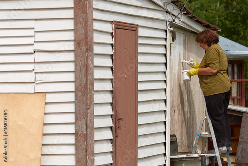A middle-aged woman in  T-shirt outdoors paints a wooden building with a brush with white paint © gogiya