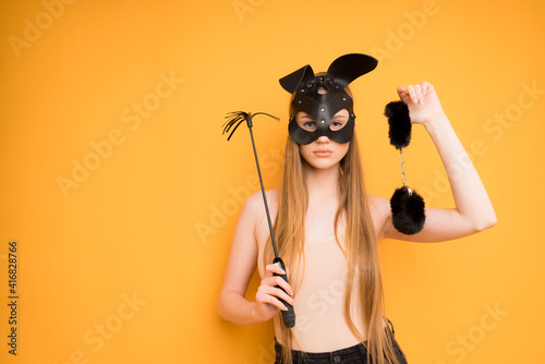 Sexy woman in a leather black fetish mask of a hare with a whip in her hands and handcuffs for sex. Stands on a yellow background