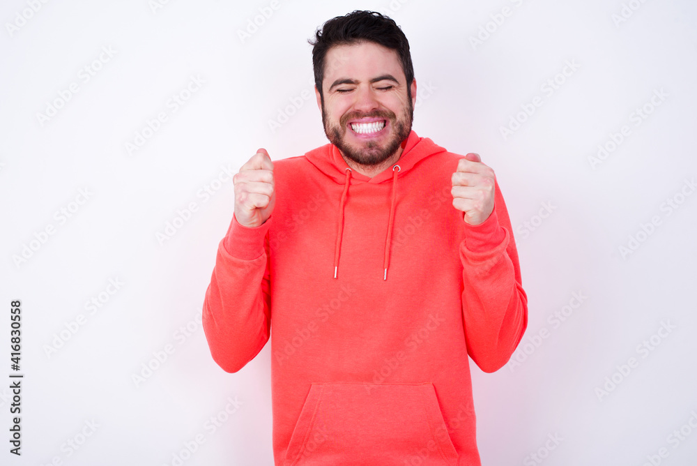 young Caucasian bearded man wearing pink hoodie against white background rejoicing his success and victory clenching fists with joy being happy to achieve aim and goals. Positive emotions, feelings. young Caucasian bearded man wearing pink hoodie against white background rejoicing his success and victory clenching fists with joy being happy to achieve aim and goals. Positive emotions, feelings.