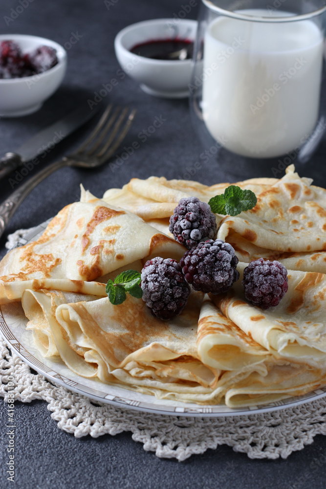 Thin pancakes with wheat flour, eggs and kefir, served with blackberries, jam and a glass of milk on dark gray table Thin pancakes with wheat flour, eggs and kefir, served with blackberries, jam and a glass of milk on dark gray table