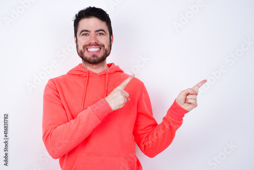 Positive young Caucasian bearded man wearing pink hoodie against white wall with beaming smile pointing with two fingers and looking on empty copy space. Advertisement concept.