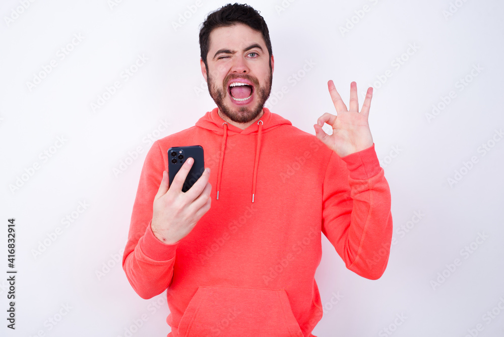 Happy young Caucasian bearded man wearing pink hoodie holding telephone against white background sending a message on his smartphone or taking a selfie and making ok sign with his hand. Happy young Caucasian bearded man wearing pink hoodie holding telephone against white background sending a message on his smartphone or taking a selfie and making ok sign with his hand.