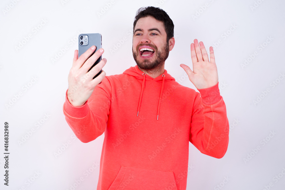 Portrait of happy friendly young Caucasian bearded man wearing pink hoodie holding telephone against white background taking selfie and waving hand, communicating on video call, online chatting. Portrait of happy friendly young Caucasian bearded man wearing pink hoodie holding telephone against white background taking selfie and waving hand, communicating on video call, online chatting.