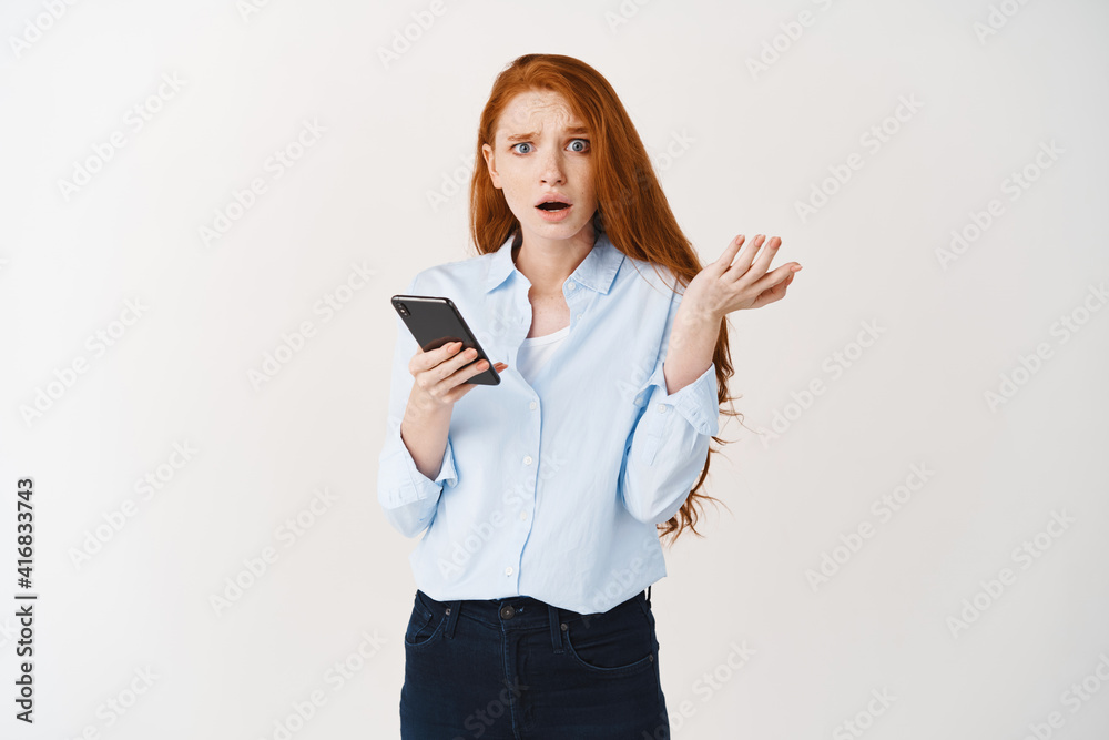 Confused and worried redhead woman receiving bad news on phone, looking at camera puzzled and shrugging, dont know what happening, app crash, white background Confused and worried redhead woman receiving bad news on phone, looking at camera puzzled and shrugging, dont know what happening, app crash, white background
