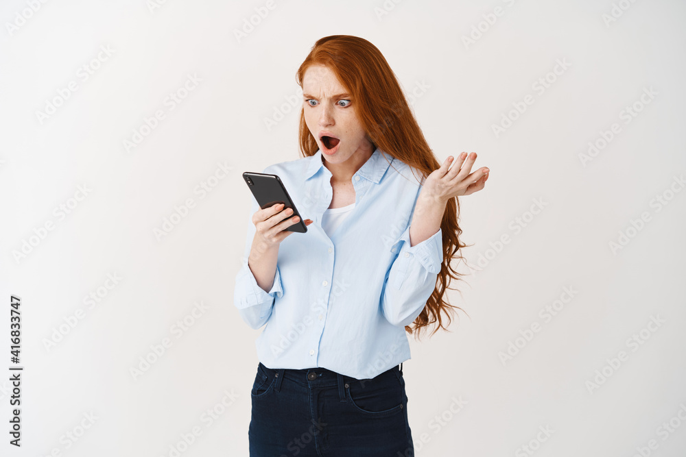 Shocked and angry woman with red hair receive spam on phone, reading irritating message on smartphone and looking outraged, white background Shocked and angry woman with red hair receive spam on phone, reading irritating message on smartphone and looking outraged, white background