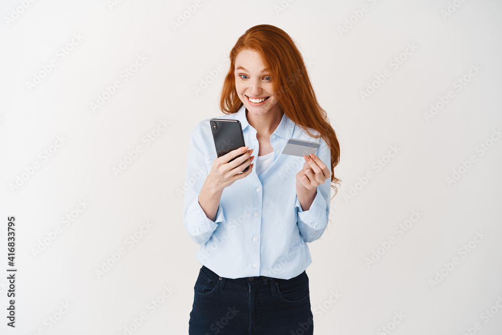 Redhead woman making online order, holding plastic credit card and looking at smartphone screen surprised, white background Redhead woman making online order, holding plastic credit card and looking at smartphone screen surprised, white background