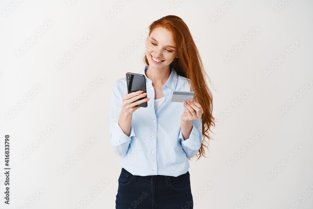 Smiling girl shopping online, holding credit card and making purchase on smartphone, white background Smiling girl shopping online, holding credit card and making purchase on smartphone, white background