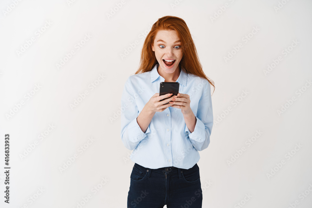 Happy redhead woman scream from joy and surprise as reading promotion deal on smartphone screen, smiling amazed, standing over white background Happy redhead woman scream from joy and surprise as reading promotion deal on smartphone screen, smiling amazed, standing over white background