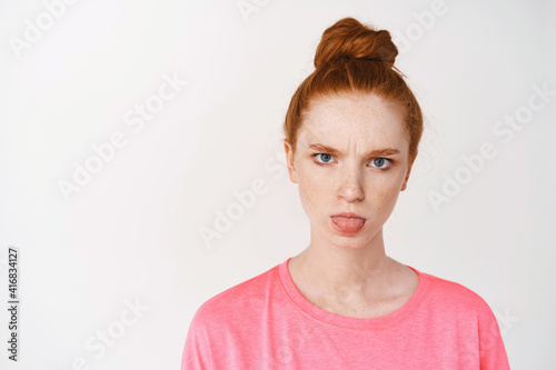 Close-up of silly redhead teen girl showing tongue and frowning, standing against white background angry