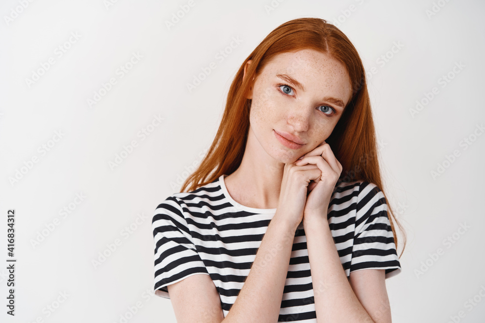 Lovely girl with ginger hair and blue eyes looking with admiration and care at camera, leaning face on hands and daydreaming, standing over white background Lovely girl with ginger hair and blue eyes looking with admiration and care at camera, leaning face on hands and daydreaming, standing over white background