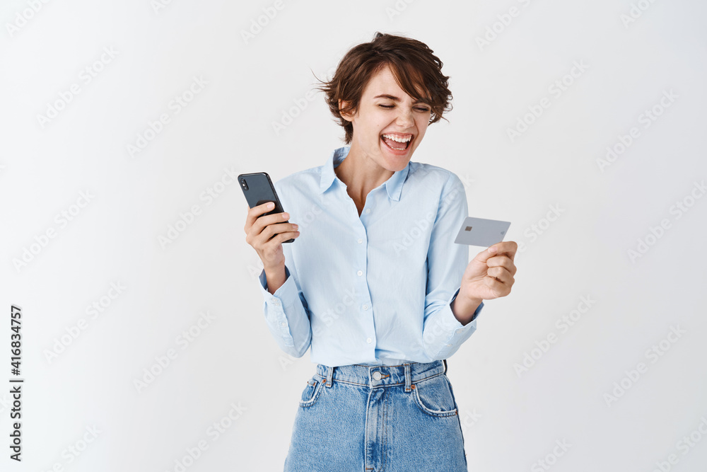 Naklejka premium Cheerful laughing woman looking at plastic credit card and using smartphone, order online, shopping in internet, standing against white background