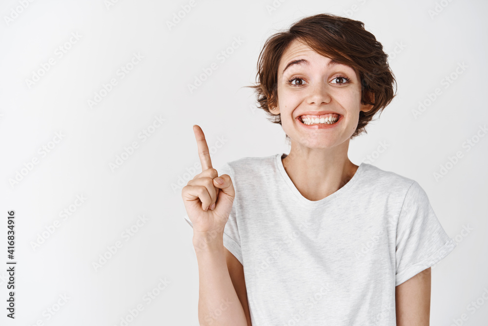 Close up portrait of happy smiling woman without makeup, wearing t-shirt and pointing up, showing logo, standing against white background Close up portrait of happy smiling woman without makeup, wearing t-shirt and pointing up, showing logo, standing against white background