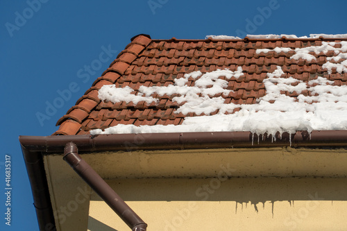 A house with orange walls and a red tiled roof. The roof of the building is covered with snow and ice after a snowfall. Hanging and falling icicles are a danger to people.