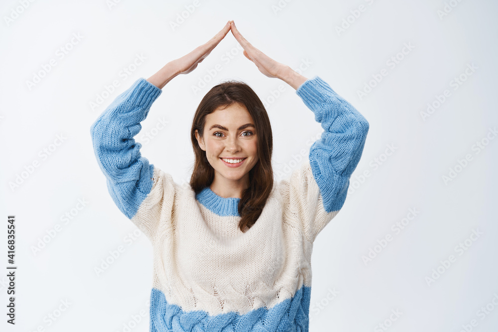 Insurance and real estate concept. Portrait of smiling woman in sweater showing home roof, making rooftop hands and looking joyful at camera, white background