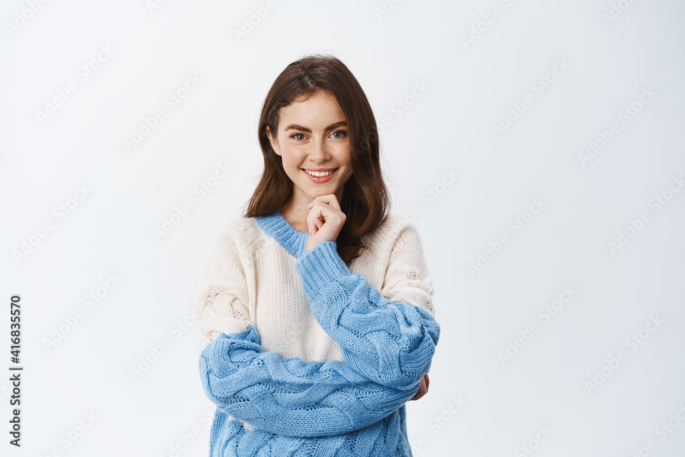 Determined woman in sweater looking thoughtful and pleased at camera, smiling with pensive face, standing against white background Determined woman in sweater looking thoughtful and pleased at camera, smiling with pensive face, standing against white background