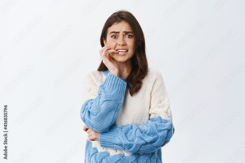 Scared and anxious woman standing worried and nervous, biting finger nail and frowning, trembling terrified, standing in sweater against white background Scared and anxious woman standing worried and nervous, biting finger nail and frowning, trembling terrified, standing in sweater against white background