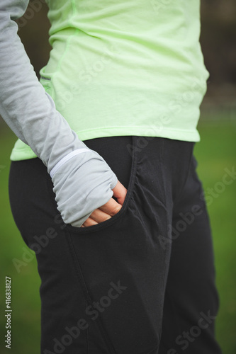 Side view of young fitness woman in sportswear holding her hand in her pocket. A girl stands outdoors in black pants and a light green sweater. Healthy lifestyle