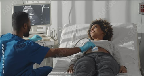 Young male african doctor, looking at screen performing ultrasound scan of pregnant woman