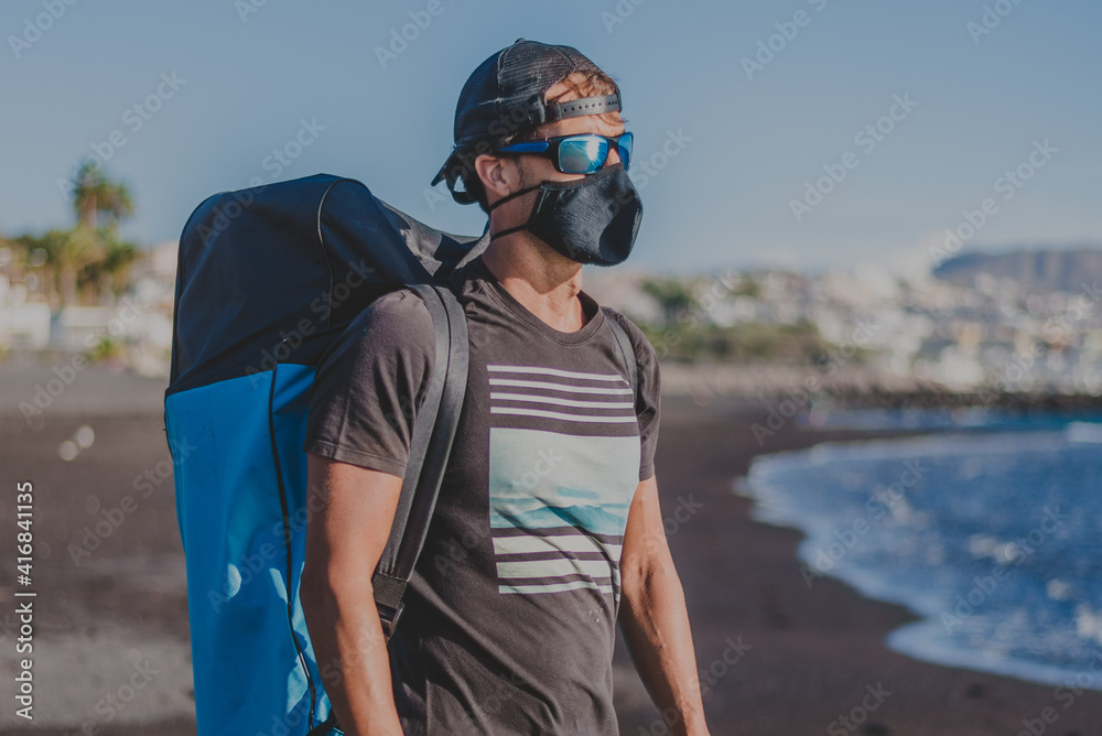 young man in cap and glasses and surf clothes looks at the sea with mask corona