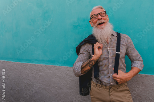 an older man in hipster clothes and glasses and a long white beard poses on a blue wall focus on head with a big smile