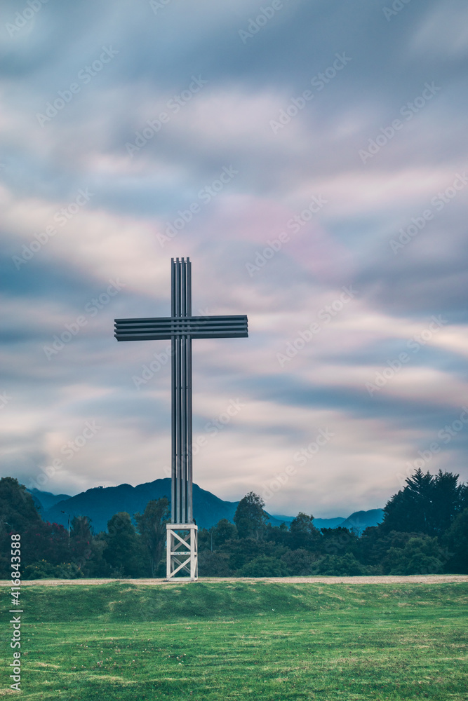 Huge cross in a park with some green trees, on a cloudy day and some hills in the background 