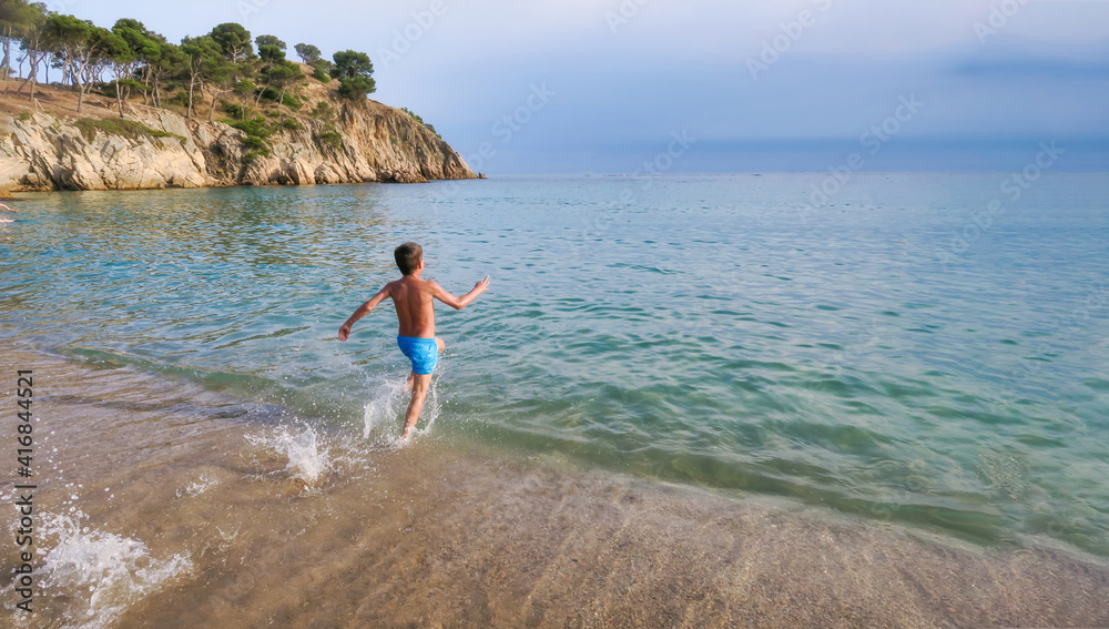 child runs along the shore towards the sea splashing water to take a dip in a beach of crystal clear turquoise waters with a cliff in the background