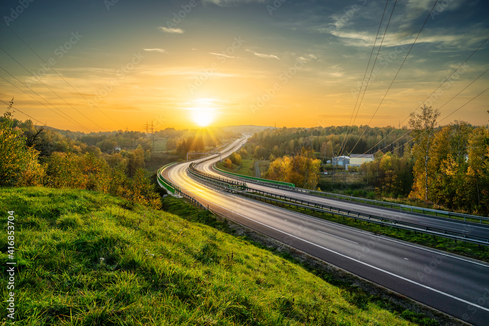 Empty asphalt highway winding through forested landscape in autumn colors at sunset