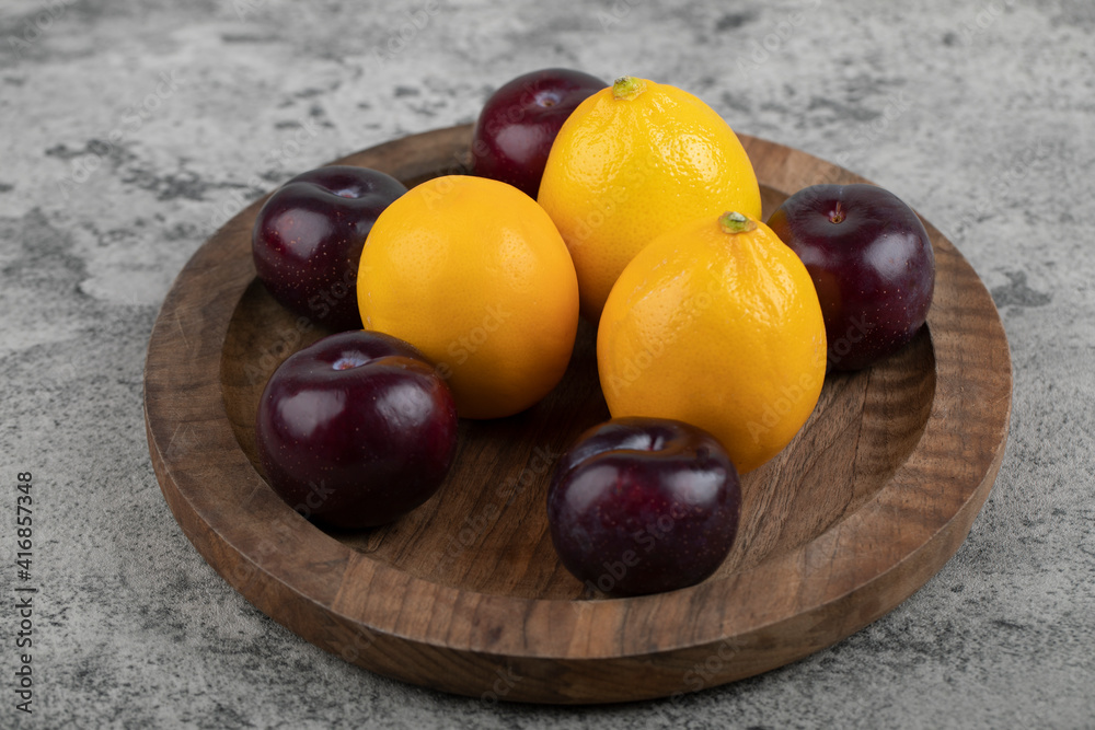 A wooden plate of purple plums and fresh lemons on stone background A wooden plate of purple plums and fresh lemons on stone background