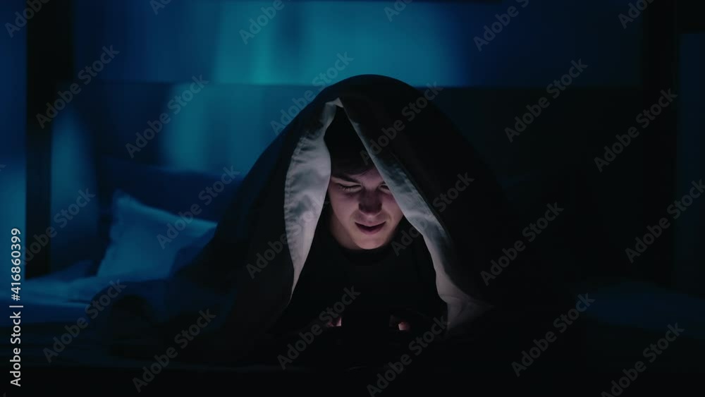 Night chat. Mobile communication. Social media addiction. Curious guy enjoying using phone in bed under blanket late at home in dark blue light. Night chat. Mobile communication. Social media addiction. Curious guy enjoying using phone in bed under blanket late at home in dark blue light.