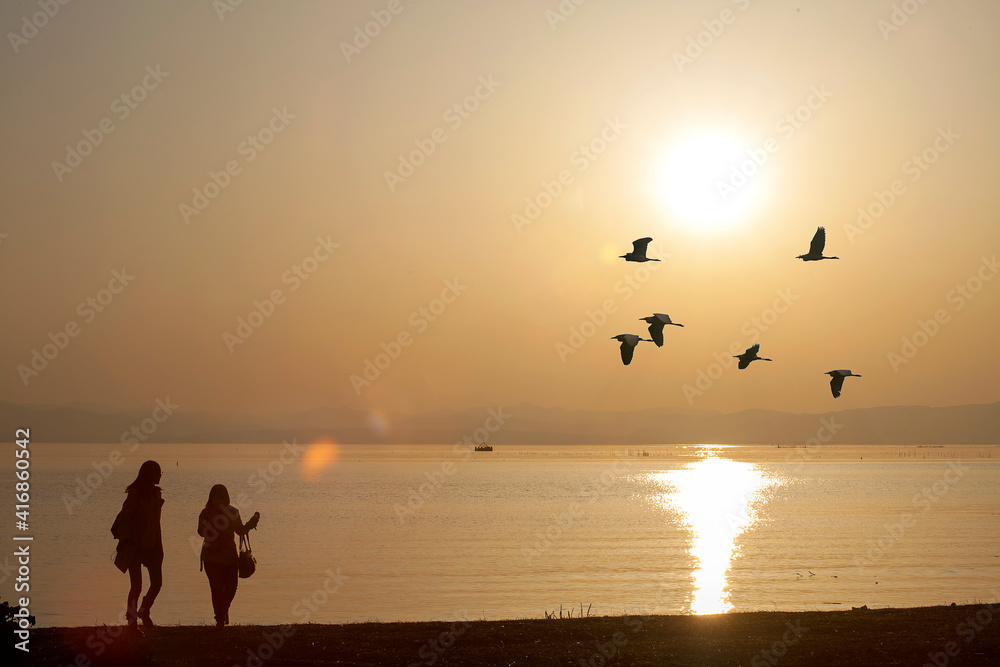 two person standing beside the  lake with bird fly in the sky druning sunset