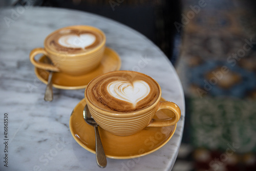 Two yellow cups of hot cappuccino on marble table background. Heart shape as art latte for love