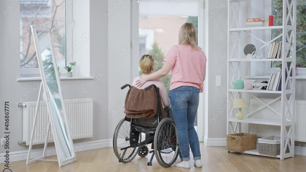 Back view wide shot of positive disabled woman on wheelchair talking with friend smiling. Happy Caucasian paralyzed invalid gossiping with young lady at home indoors. Friendship and illness. Back view wide shot of positive disabled woman on wheelchair talking with friend smiling. Happy Caucasian paralyzed invalid gossiping with young lady at home indoors. Friendship and illness.