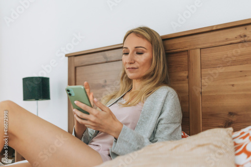 Beautiful blonde young woman in home wear using mobile on bed in bedroom photo