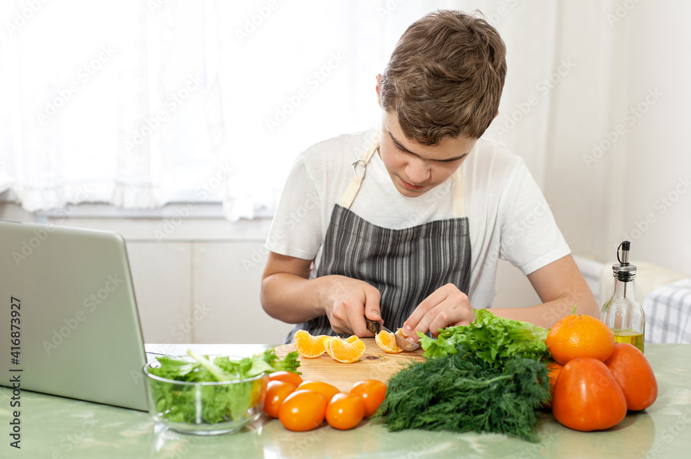 A teenage boy prepares a fruit salad on a recipe from the Internet. The boy cuts lettuce on the kitchen table, in front of him is a laptop. The child is engaged in household chores. A teenage boy prepares a fruit salad on a recipe from the Internet. The boy cuts lettuce on the kitchen table, in front of him is a laptop. The child is engaged in household chores.