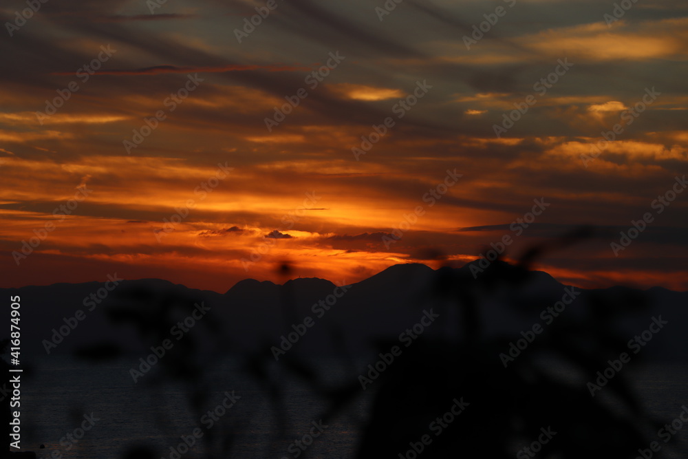 江ノ島で夕方に木の葉の間から望む伊豆半島シルエットとオレンジ色の夕日