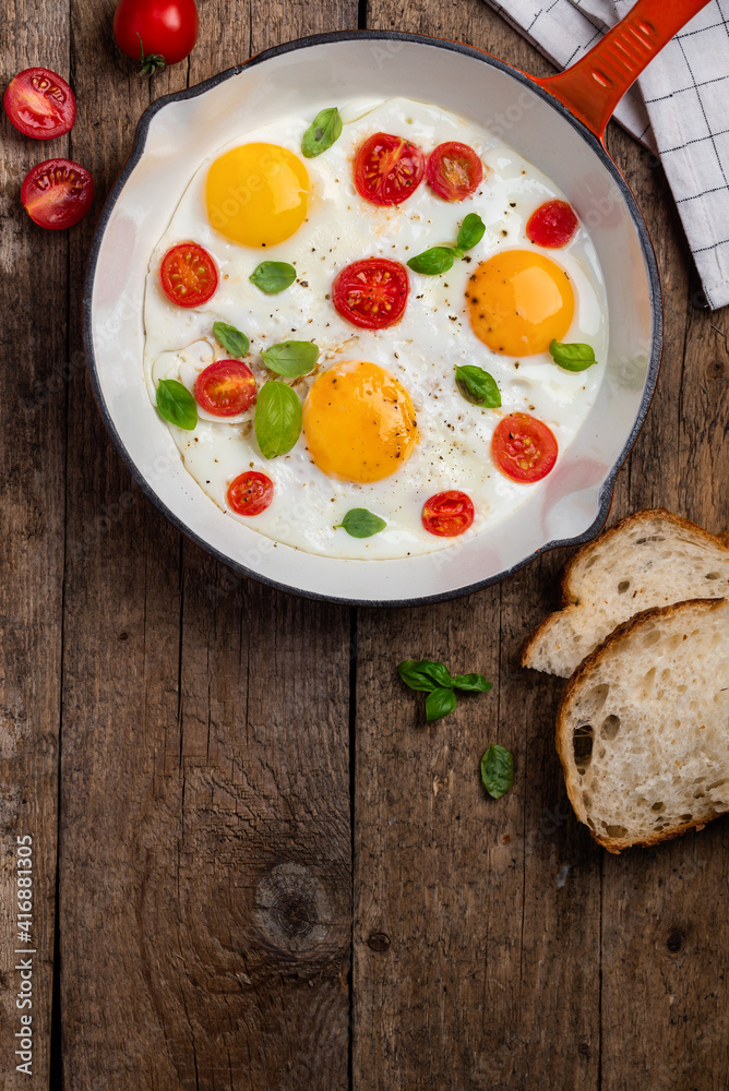 Fried eggs with cherry tomatoes and basil in a red pan for breakfast. Wood background. Copy space Fried eggs with cherry tomatoes and basil in a red pan for breakfast. Wood background. Copy space