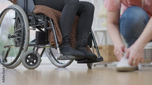 Unrecognizable paraplegic woman sitting in wheelchair with blurred caregiver preparing shoes to put on. Kind Caucasian friend or nurse taking care of disabled person indoors.