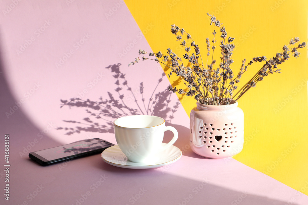 A bouquet of lavender with a cup of tea and a phone on a yellow-pastel background in the sun with a shadow from objects, side view A bouquet of lavender with a cup of tea and a phone on a yellow-pastel background in the sun with a shadow from objects, side view
