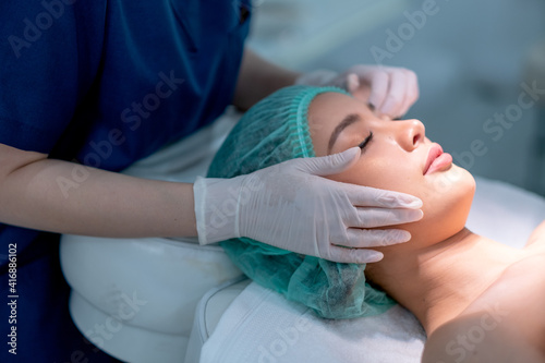 Beauty clinic hands touch on face of customer woman who lie on bed with light during process of treatment.