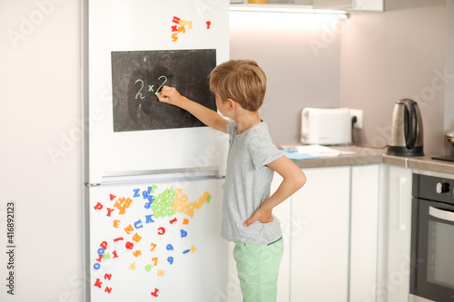 A 8-year-old schoolboy writes white chalk on a magnetic black board attached to a fridge.