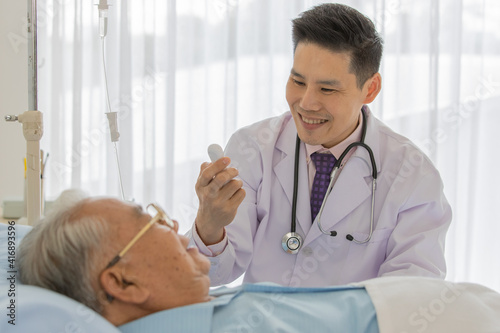 A young handsome asian male doctor wearing white lab coat with stethoscope checking temperature from thermometer in his hand smiling nearby old fat asian patient with gray hair wearing eyeglasses 