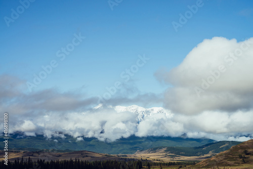 Awesome alpine landscape with great snowy mountain range in big low clouds under blue sky. Scenic view to snowy ridge above forest in large low cloud. Colorful mountain scenery with high mountains.