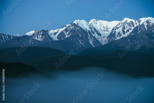 Fototapeta Naklejka Na Ścianę i Meble -  Atmospheric mountains landscape with dense fog and great snow mountain top under twilight sky. Alpine scenery with big snowy mountains over thick fog in night. High snow pinnacle above clouds in dusk.